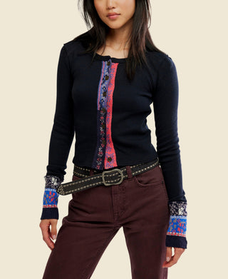 A woman wearing a black long-sleeve knit noa cardigan from free people with a colorful vertical patterned placket and matching patterned cuff trim, paired with dark brown pants and a studded belt, against a plain white background.