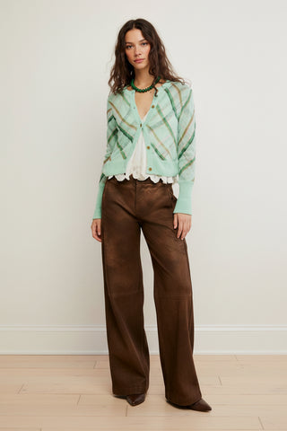 A woman wearing a mint green knit cardigan, the Free People Highland Mist Cardi, with diagonal plaid stripes in olive green and tan, styled over a white ruffled blouse and brown pants.