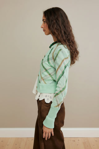 A woman wearing a mint green knit cardigan, the Free People Highland Mist Cardi, with diagonal plaid stripes in olive green and tan, styled over a white ruffled blouse and brown pants.