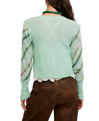 A woman wearing a mint green knit cardigan, the Free People Highland Mist Cardi, with diagonal plaid stripes in olive green and tan, styled over a white ruffled blouse and brown pants.